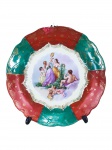Oscar Schlegelmilch (Alemanha, Langewiesen), um (1) prato decorativo confeccionado em porcelana alemã na tonalidade branca, centro representando cena ninfa, borda bicolor nas tonalidades vermelha e verde com decoração floral em ouro, apresenta bicado com restauro manual na borda e falhas na pintura, suporte de brinde com colado, medindo 29cm.