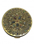 METAL DOURADO, uma (1) caixa porta pó confeccionada em metal dourado, tampa com rico cinzelado de inspiração floral e arabescos, aplicação de pedras coloridas em tons de azul, verde, vermelho e lilás, interior espelhado, sinais de uso e leves oxidações medindo 8cm de diâmetro.