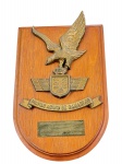 COLECIONISMO, uma (1) placa comemorativa de parede, confeccionada em madeira envernizada, com aplicação em metal dourado/patinado representando águia em voo sobre brasão do Mando Aéreo de Canarias, com faixa inferior em relevo. Possui placa metálica gravada com dedicatória Del Mando Aéreo de Canarias a la Fragata de la Armada Brasileña Defensora  Las Palmas, Marzo de 1989. Medida : 28 x 18 cm Peça com sinais de uso.