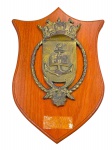 COLECIONISMO, Uma (1) placa comemorativa de parede, em formato de escudo, confeccionada em madeira envernizada, com aplicação em metal dourado/patinado representando brasão naval com âncora, livro aberto, cordame e elementos em relevo na parte superior. Não possui placa inferior . Medida 39 x 27 cm