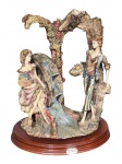 ESCULTURA, um (1) grupo escultórico confeccionado em resina italiana representando um casal em um ambiente pastoral com um moinho de água, apoiado sobre base oval em madeira escalonada, medindo 39x33x24cm.