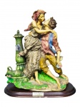ESCULTURA, um (1) grupo escultórico confeccionado em resina italiana representando um  casal romântico abraçado junto a um antigo chafariz, esmaltado em policromia, apoiado sobre base oval em madeira escalonada, medindo 33,5x30x15cm, base apresentando leves falhas.
