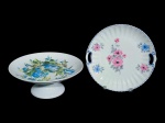 PORCELANA - Lote com duas (2) peças confeccionadas em porcelana  , sendo: A) um (1) prato para bolo da manufatura Real, com decoração floral policromada, borda canelada, filete azul e alça vazada lateral , medida : 26 cm ; B) uma (1) fruteira da manufatura Schmidt, com base elevada, formato circular e decoração floral em tons de azul, verde e amarelo. Medida : 11 x 27 cm . Prato apresenta fio de cabelo .