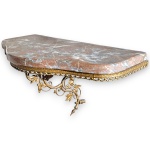 CONSOLE SUSPENSA EXECUTADA EM AÇO PATINADO, COR OURO ENVELHECIDO, RICO TRABALHO ORNAMENTAL EM VOLUTAS, FOLHAGENS E ACABAMENTO VAZADO DE INSPIRAÇÃO ROCOCÓ, MEDINDO 30 X 51 X 27 CM, ACOMPANHADO DE TAMPO EM MÁRMORE ROSADO VEIADO, FORMATO RECORTADO. APRESENTA MARCAS DO TEMPO, USO, DESGASTES E/OU PEQUENAS IMPERFEIÇÕES COMPATÍVEIS COM SUA UTILIZAÇÃO E ESTADO DE CONSERVAÇÃO, CONFORME FOTOS EXTRAS. INFORMAMOS AINDA QUE NÃO REALIZAMOS EMBALAGEM DE MÓVEIS, SENDO A RETIRADA, ACONDICIONAMENTO, PROTEÇÃO E TRANSPORTE DE TOTAL RESPONSABILIDADE DO ARREMATANTE E/OU TRANSPORTADOR CONTRATADO.