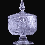 BONBONNIÈRE SOBRE PEDESTAL EXECUTADA EM VIDRO TRANSLÚCIDO MOLDADO E LAPIDADO, COM CORPO DE FORMATO CILÍNDRICO DECORADO POR GRANDES MEDALHÕES OVALADOS COM MOTIVOS DE ESTRELAS EM ROSETAS E LAPIDAÇÕES GEOMÉTRICAS EM RELEVO, ASSENTADA SOBRE HASTE CURTA COM CABOCHÃO E BASE CIRCULAR LISA, ACOMPANHADA DE SUA RESPECTIVA TAMPA ENCIMADA POR PEGA PONTIAGUDA BALAÚSTRE MULTIFACETADA, MEDINDO 23,5 X 14,5 CM. APRESENTA MARCAS DO TEMPO, USO, DESGASTES E/OU IMPERFEIÇÕES COMPATÍVEIS COM SUA UTILIZAÇÃO E ESTADO DE CONSERVAÇÃO, CONFORME FOTOS EXTRAS. ESCLAREÇA TODAS AS DÚVIDAS ANTES DE EFETUAR SEU LANCE, QUE SERÁ CONSIDERADO IRREVOGÁVEL E IRRETRATÁVEL. CONSULTE PREVIAMENTE OS TERMOS E CONDIÇÕES DISPONÍVEIS NO SITE. CONTATO (21) 98264-3000.