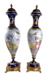 SÉVRES - PAR DE IMPORTANTES ÂNFORAS EM PORCELANA DE SÈVRES, "BLEU DE ROI" FORMATO BALAÚSTRE ALONGADO  APRESENTANDO EXCEPCIONAL TRABALHO DE PINTURA MANUAL EM RESERVAS CENTRAIS COM CENAS ROMÂNTICAS EM PAISAGEM NATURALISTA, ORNAMENTAÇÃO EM OURO BRUNIDO COM MOTIVOS FLORAIS E ARABESCOS NO GARGALO E NA BASE,  ASSENTES SOBRE BASES QUADRANGULARES EM BRONZE DOURADO COM PÉS EM VOLUTAS MANTENDO A IMPONÊNCIA. PEÇA EM ÓTIMO ESTADO DE CONSERVAÇÃO. MEDIDAS: 65 CM DE ALTURA
