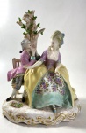 MEISSEN - GRUPO ESCULTÓRICO EM POCELANA DE MEISSEN REPRESENTANDO CENA GALANTE EM UM JARDIM SOB UMA ÁRVORE QUE CONTÉM UM QUERUBIM ACIMA DO CASAL SIMBOLIZANDO O AMOR. MEDIDAS: 30 CM DE ALT.  X 24 CM DE LARG.