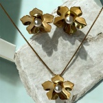 SEMI JÓIAS - Conjunto de semi joias banhadas a ouro com design floral, composto por colar com pingente de flor e brincos coordenados, destacando pérola central. Peças elegantes com acabamento polido e estilo clássico.