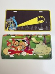 OBJETOS DECORATIVOS - Lote com 2 placas decorativas, sendo uma com tema Batman e outra com tema Os Flintstones. Peças em metal, medindo aproximadamente 30x15cm cada. Itens com pequenos amassados e desgastes leves.