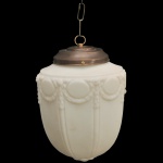 Luminária pendente de estilo Império / Art Nouveau, caracterizado por uma cúpula de vidro fosco (ou opalinado) em formato de tulipa ou pinha. O corpo de vidro apresenta decorações clássicas moldadas em alto-relevo com motivos de guirlandas e medalhões circulares na parte superior, complementados por gomos canelados verticais que descem até a base. 170 x 25 x 23 cm canopla / corrente.