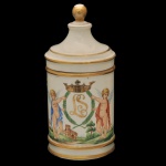Pote de farmácia em porcelana brasileira pintada a mão, produzida no Rio de Janeiro. Apresenta corpo cilíndrico de superfície branca e uma decoração em policromia com frisos a ouro e detalhes de querubins. Pega da tampa em dourado. Ass. Leonor Formenti. 21 x 11 cm.
