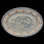 Travessa oval em porcelana chinesa com o padrão "grão de arroz", apresentando um dragão azul no centro. A peça possui decorações adicionais em tons de vermelho (rouge de fer) e elementos abstratos ou labaredas estilizadas. Marca da manufatura no verso. 30 x 22 cm.