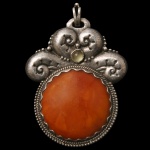 Pingente histórico em prata com uma grande gema de âmbar incrustada, ornamentado com detalhes entalhados e uma pequena pedra decorativa superior. O reverso desta joia traz uma gravação oficial Do Imperador a Imperatriz 1864 indicando que ela pertenceu a Dona Teresa Cristina, a Imperatriz do Brasil e esposa de Dom Pedro II. O desenho da peça é conhecido por refletir os modos discretos e o gosto pessoal austero da monarca.