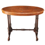 Séc. XIX - Mesa de lateral de apoio ovalada, em madeira nobre (rádica). Estilo vitoriana, conhecida em inglês como Burr Walnut Centre Table. O tampo possui um padrão espelhado (book-matched), destacando os nós naturais e os veios luxuosos da madeira nobre. Apresenta colunas duplas torneadas em cada extremidade, unidas por uma travessa central ornamentada.  A base se divide em pés no estilo cabriolet com entalhes de folhas de acanto e volutas na ponta, tipicamente equipados com rodízios de cerâmica ou latão originais da época. 73 x 90 x 59 cm.