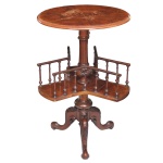 Séc. XIX - Mesa de livros giratória vitoriana em madeira nobre (nogueira) conhecida em inglês como Revolving Book Table), combina a função de uma mesa lateral ou de cabeceira com uma estante compacta para livros de uso frequente. Possui uma prateleira inferior dividida em três seções com pequenas grades vazadas de balaústres (mini colunas torneadas). Essa base gira de forma independente em torno do eixo central, facilitando o acesso aos livros sem precisar mover o móvel.  Fabricada em madeira nobre, com destaque para o acabamento do tampo que exibe os veios característicos e luxuosos da raiz de nogueira. Conta com uma coluna central ricamente torneada e esculpida, apoiada sobre uma base clássica em tripé (três pés curvos) decorada com entalhes vitorianos que garantem elegância e estabilidade. 78 x 50 cm.