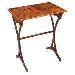 Mesa lateral de estilo Art Nouveau, marcante pela forte inspiração em formas orgânicas e da natureza. O tampo retangular apresenta um trabalho ricamente detalhado de marchetaria, combinando diferentes tonalidades e tipos de madeiras nobres para formar um desenho de folhas ornamentais, reminiscente dos designs característicos da famosa École de Nancy (como as criações do icônico designer Émile Gallé). As pernas são delgadas, esculpidas e arqueadas de forma sinuosa. Elas se unem de maneira elegante na parte inferior e são integradas por uma travessa horizontal reta esticada no centro, garantindo a estabilidade sem perder o desenho estético leve da peça. Esse mobiliário personifica a virada do século XIX para o século XX, onde o design de móveis abandonou a rigidez geométrica do passado para adotar linhas inspiradas em plantas, galhos e folhagens. 70 x 59 x 39 cm.
