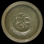 Medalhão circular em porcelana chinesa, revestido por coberta celadon de cor verde oliva. Apresenta decoração floral incisa, centralizando rosácea com dragão em leve relevo. Verso realçado por folhas alinhadas. Peça em estilo da dinastia Ming. 8 x 40 cm.