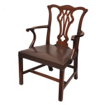 Cadeira de braços estilo Chippendale em mogno com assento estofado em couro. Apresenta o tradicional encosto vazado e recortado em formato de balaústre recortado (pierced splat), combinando linhas curvas e detalhes geométricos. Possui apoios de braço sinuosos e curvados para fora (serpentine armrests), que se conectam elegantemente às laterais da estrutura. Tipo "drop-in" (assento removível encaixado na moldura), estofado em couro marrom escuro com acabamento fixado por tachas metálicas decorativas nas bordas. Pernas frontais retas e quadradas unidas por travessas em formato de "H", conferindo alta estabilidade estrutural típica dos modelos utilitários e residenciais do período Georgiano. 97 x 68 x 50 cm.