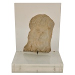 Escultura em pedra calcária natural, mantendo texturas rústicas e marcas de entalhe características do artista. Exibe um rosto masculino barbado e expressivo, frequentemente associado a figuras bíblicas, anjos ou santos em sua produção artística. O fragmento está fixado por um pino de metal em uma base expositora moderna de acrílico transparente.