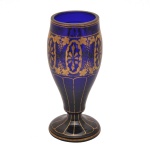Cálice decorativo vintage, em vidro de cor azul-cobalto e ricamente ornamentado com detalhes em ouro. Corpo alongado e facetado com uma faixa central dourada contendo arabescos e padrões florais clássicos gravados. O corpo se estreita até uma haste fina, conectando-se a uma base circular firme com acabamento também em filete dourado.
