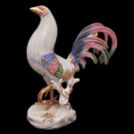 Portugal. Vista Alegre - Estatueta retratando um galo em porcelana caracterizada por pintura policromada em tons de azul, verde e rosa, além de detalhes dourados, posicionada sobre uma base naturalista decorada. Marca da manufatura Vista Alegre, no verso. Pintado à mão. 38.5 cm. Apresenta restauro no rabo. 38 x 30 cm.
