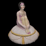 Porta-joias em fina porcelana esmaltada com detalhes ricamente pintados a mão, de estilo Art Déco, Fabricada na Alemanha por volta das décadas de 1920 e 1930. Tampa exibe uma escultura detalhada de uma jovem melindrosa (flapper girl) sentada sobre um travesseiro macio, que funciona como a tampa removível do pote. Apresenta os traços clássicos das Annes Folles (os Anos Loucos), incluindo o icônico corte de cabelo curto estilo bob preto, batom vermelho marcante e uma faixa de cabelo colorida na testa. 25 x 20 cm. Restauro na parte de baixo.