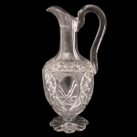 BACCARAT - Belíssimo decanter em cristal lapidado (frequentemente chamada de claret jug ou licoreira), um item clássico que remonta ao século XIX ou início do século XX. Corpo oval e bojudo, pescoço longo facetado e bico curvo pronunciado. Apresenta intrincados padrões geométricos em relevo, incluindo lapidações em formato de losangos cruzados cortes no estilo diamante e motivos de folhas. Alça curva e lisa acoplada manualmente e uma sofisticada base circular trabalhada em formato de roseta ou estrela lapidada. 30 x 13 cm.