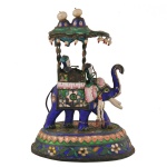 Estatueta indiana vintage de elefante real com palanquim (howdah) e figuras, tradicionalmente confeccionada em prata com esmaltes cloisonné, Representa um elefante de procissão real ricamente ornamentado, transportando um condutor (mahout) à frente e uma figura de prestígio (frequentemente um Marajá) dentro do palanquim coberto por um dossel decorativo. A base e o topo costumam apresentar pingentes decorativos em imitação de pérolas ou pequenas contas. Esta peça utiliza a arte tradicional indiana conhecida como Meenakari (esmaltação sobre metal), muito associada a centros artesanais como Jaipur, Lucknow e Varanasi. 18 x 13 x 9 cm.