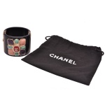 CHANEL. Bijoux  - Bracelete de grife autêntico da Chanel, conhecido especificamente como Chanel Black Resin Multicolor Stone Gripoix Cuff Bracelet. Confeccionado em resina preta brilhante de alta espessura. Apresenta um medalhão central em formato de cruz de malta ou flor. Ele é adornado com pedras esmaltadas coloridas em tons de rosa, roxo e verde texturizado (estilo vidro Gripoix). O icônico fecho duplo CC dourado da marca fica posicionado em destaque no centro da peça. Acompanha Dust bag em tecido para guardar e proteger a peça contra riscos, poeira e danos ao longo do tempo. A tipografia "CHANEL" é limpa, centralizada e utiliza a fonte padrão geométrica registrada da marca. 5 x 6 cm. Peça nova, sem uso.
