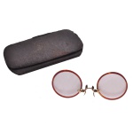 Óculos (pince-nez) vintage redondos com armação dourada e lentes colorida, possivelmente do início do século XX, Acompanha estojo apresentando um acabamento texturizado escuro, semelhante a couro ou material rígido.