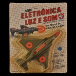 COLECIONISMO - Caça supersônico da série "Eletrônica Luz e Som" da marca Estrela, lançado nos anos 90. Embalagem original de fábrica (lacrado), com gráficos em grade e o avião posicionado centralmente. Possui características eletrônicas, como luzes que piscam no nariz do avião e sons de decolagem ou metralhadora quando o botão é pressionado. Por ser um item antigo e lacrado, é considerado um item de colecionador, embora a funcionalidade eletrônica não possa ser garantida devido à idade das pilhas originais.