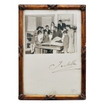 Fotografia Histórica e Autografada do ex-presidente brasileiro Juscelino Kubitschek reunido com crianças e jovens, datada de 1956, ano em que ele assumiu a presidência da República. No registro, o político aparece ao centro, de terno, folheando um grande livro ou álbum cercado por jovens atentos. Abaixo da imagem em preto e branco, nota-se a assinatura oficial estilizada feita de próprio punho ("J. Kubitschek") e a marcação do ano "1956" no canto inferior direito. O item está preservado em uma moldura de madeira trabalhada com pátina dourada. Ass. 17 x 11 cm (MI). 19 x 13 cm (ME).
