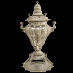 Tea Kettle em prata chinesa com sua bandeja, profusamente decorada por adornos fitomórficos e elementos estilizados. Bojo piriforme, moldado com gomos e ornamentado por figuras variadas como casais, musicistas e letrados em reservas. Apresenta alças laterais reviradas e pequena torneira à frente com pega em arabesco. Gargalo retraído e bordado por perolado, encimado por segmento cônico montado em degraus. Base circular realçada por vazados, apoiada em peanha quadrangular com pés revirados e garras. Presentoir de formato triangular com laterais recurvas, acompanhando o estilo. Apresenta contrastes com escritas e marca.