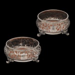 Elegante par de suportes para garrafa (também conhecidos como wine coasters, confeccionados em prata de lei decorada. Os suportes exibem um requintado trabalho de prataria vazada e cinzelada em relevo, com motivos clássicos de folhagens e volutas. Cada peça possui uma cartela central polida (para gravação de iniciais ou brasão) e é sustentada por pés ornamentados em formato de garras ou volutas.  Iniciais entrelaçadas em reserva central.