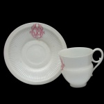 Xícara com seu pires em porcelana francesa branca, moldados com finos gomos. Apresentam monogramas de cor magenta. Bojo com base cônica, encimado por gargalo reto e alça em voluta. Marca desconhecida sob a base. 6 x 8 cm (xícara) / 12 cm (pires).