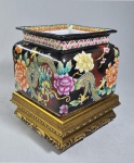Cachepot quadrado em porcelana sem marca, bordas chanfradas, decorado com dragão e elementos orientais sobre fundo negro. Base em madeira dourada. Medida total: 22.5 cm de altura.