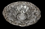 Antiga fruteira em metal espessurado a prata, com corpo circular ricamente trabalhado em relevo, apresentando ornamentação em volutas, elementos florais e bordas recortadas. Possui centro circular liso, em contraste com a decoração em estilo clássico. Medidas: 20 x 7 cm.
