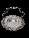 Antiga fruteira em metal espessurado a prata, com corpo circular ricamente ornamentado em relevo, apresentando bordas recortadas com motivos florais e volutas. Possui alça superior elevada em desenho sinuoso. Medidas: 20 x 17,5 x 13 cm.
