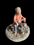 Escultura em biscuit policromada representando camponês sentado sobre base naturalista, segurando pequeno cesto, com detalhes em relevo e rica vestes e expressão facial. Base com marca Made in Portugal. Medidas: 21 x 23,5 x 21 cm.