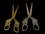 Duas tesouras, sendo uma em metal espessurado a prata e outra dourada, com cabos ricamente ornamentados em relevo com motivos florais e vegetalistas, apresentando lâminas afiladas e acabamento decorativo. Medidas: 14,5 x 6,5 cm.
