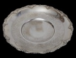 Fruteira em metal espessurado a prata, com borda recortada e ricamente ornamentada em relevos de inspiração rococó, apresentando centro com delicada decoração gravada. Medidas: 7 x 35 cm.