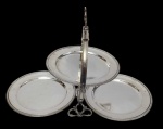 Antiga petisqueira dobrável de três bandejas, produzida em metal espessurado a prata, com estrutura articulada, pés ornamentados e gravação decorativa central em arabescos no espelho. Medidas: 43 x 28 cm.