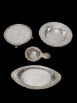 Lote contendo 4 peças diversas, produzidas em metal espessurado a prata, sendo 1 salva redonda com pés, 1 porta garrafas, 1 travessa em formato navete e 1 coador de chá. Medidas: 18 x 3 cm / 15 x 3 cm / 21 x 10 x 2 cm / 12 x 2 x 8 cm.