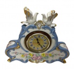 Relógio de mesa em porcelana na cor branca e azul, rosas em relevo, duas esculturas ao topo no feitio de pombinhos, relógio  com funcionamento a pilha. Medindo:  22 x 41 cm. Uma das asas apresenta bicado.