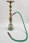 Elegante narguilé composto por reservatório em vidro decorado com pintura floral policromada, corpo em metal dourado torneado e 2 mangueiras revestidas em tecido com detalhes coloridos. Medindo 56 cm de altura.