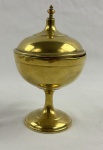 Pote com tampa em metal dourado, de corpo bojudo apoiado sobre haste central e base circular, apresentando linhas sóbrias e acabamento polido. Tampa removível encimada por pináculo torneado. Medindo 16 cm.