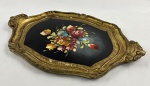 Bandeja vintage de formato oval recortado, confeccionada em madeira, Itália (estilo florentino / veneziano). Fundo central em laca preta fosca, adornado com exuberante buquê floral pintado à mão em rica policromia. Borda ricamente trabalhada com volutas, relevos e arabescos barrocos, exibindo acabamento em folha de ouro dourada. Apresenta duas alças integradas nas extremidades. Medindo: 43,5 x 28 cm.