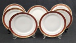 Sete pratos rasos em porcelana polonesa Karolina, na cor branca esmaltada, com abas em tom rubi decoradas por arabescos e folhagens em ouro, bordas filetadas. Medida: 24,5 cm. Apresenta marcas de desgaste na ornamentação em ouro.