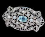 Broche Art Déco em prata com vazados, folhagens e lírios. Cabochon central em pedra azul de 1 crt, 2 diamantes lapidação brilhante de 0,20 crt e ornamentação com diversos pequenos brilhantes. Med.: 4,5 x 2,5cm. Peso total bruto: 6.5g