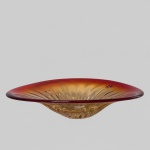 Centro de mesa circular em vidro artístico de parede espessa, apresentando gradação cromática do âmbar ao vermelho-rubi. Interior com lapidação radial em forma de raios e decoração pontual de estrelas de oito pontas, executadas por gravação. Medindo: